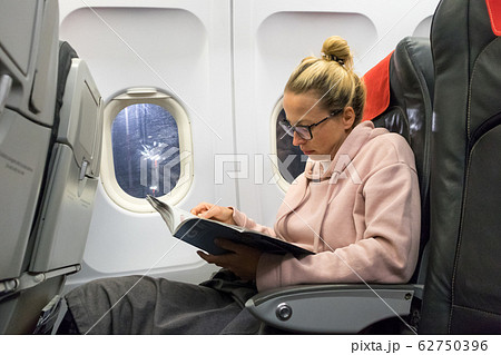 Casual young woman reading magazine and listening to music on airplane. Female traveler reading seated in passanger cabin. 62750396