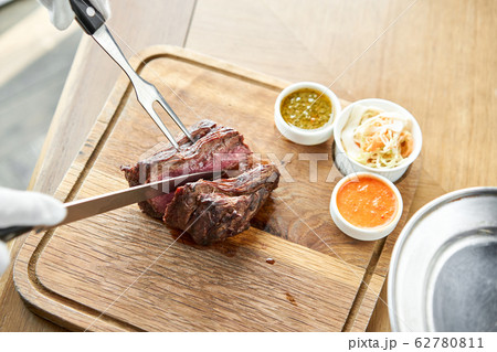The waiter is butchering griled meat with a knife. Grilled fillet steaks with Pickled cabbage and two sauces. Serving on a wooden Board. Barbecue restaurant menu. The waiter is butchering griled meat with a knife. Grilled fillet steaks with Pickled cabbage and two sauces. Serving on a wooden Board. Barbecue restaurant menu. 62780811