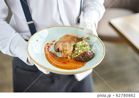 A waiter in a white shirt and apron holds a ready meal in the restaurant.Plate Duck leg su-vide, with bulgur and onion marmalade. 62780862