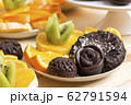 Delicious vegan snack. Chocolate candies and cookies sprinkled with coconut flakes lie with chopped fruits on a white saucer on a wooden table. Homemade baking with health benefits. Healthy meal 62791594