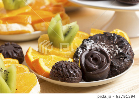 Delicious vegan snack. Chocolate candies and cookies sprinkled with coconut flakes lie with chopped fruits on a white saucer on a wooden table. Homemade baking with health benefits. Healthy meal Delicious vegan snack. Chocolate candies and cookies sprinkled with coconut flakes lie with chopped fruits on a white saucer on a wooden table. Homemade baking with health benefits. Healthy meal 62791594