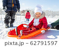 Winter activities for children. A little girl in a red coat sits on an orange sled before descending from a snow slide. Behind her are other children and a school in the distance. Snowy weather 62791597