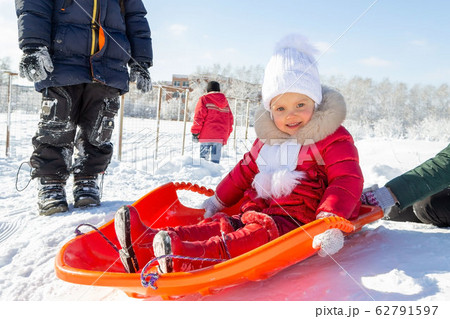 Winter activities for children. A little girl in a red coat sits on an orange sled before descending from a snow slide. Behind her are other children and a school in the distance. Snowy weather 62791597