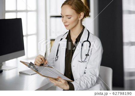 Cheerful smiling female doctor using clipboard in clinic. Portrait of friendly physician woman at work place. Perfect medical service in hospital. Medicine concept 62798190