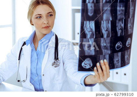 Doctor woman examining x-ray picture near window in hospital. Surgeon or orthopedist at work. Medicine and healthcare concept. Blue colored blouse of a therapist looks good Doctor woman examining x-ray picture near window in hospital. Surgeon or orthopedist at work. Medicine and healthcare concept. Blue colored blouse of a therapist looks good 62799310