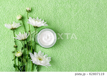 Facial cream in an open glass jar and white chrysanthemum flowers on a green terry towel Facial cream in an open glass jar and white chrysanthemum flowers on a green terry towel 62807119
