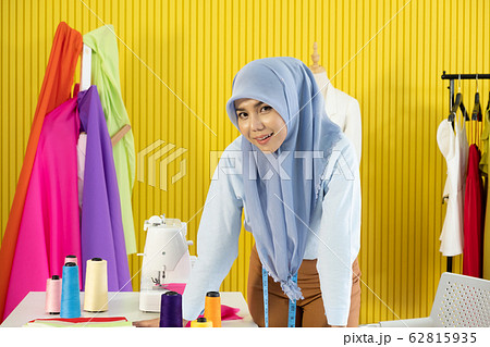 A young beautiful Asian Muslim woman designer who wore a hijab as a startup business owner standing in her working studio room with colorful cloth, dress, thread and sewing machine. A young beautiful Asian Muslim woman designer who wore a hijab as a startup business owner standing in her working studio room with colorful cloth, dress, thread and sewing machine. 62815935