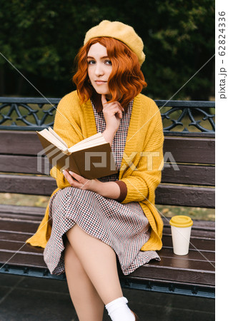 A beautiful girl with a curly hairstyle is sitting on a park bench and reading 62824336