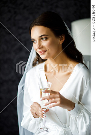 The girl sits on a soft bed, holding a glass with champagne in her hands. Wedding morning of the bride. 62826044