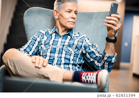 Mature male holding smartphone while resting in armchair 62831394