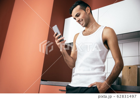 Smiling guy standing and holding smartphone at the kitchen Smiling guy standing and holding smartphone at the kitchen 62831497