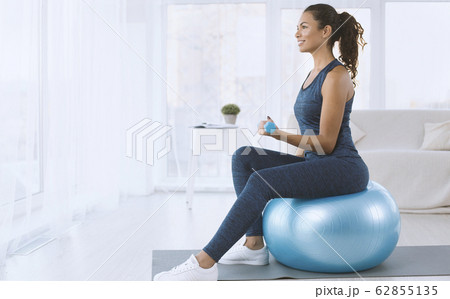Attractive Hispanic lady with dumbbells sitting on fitness ball at home, copy space 62855135