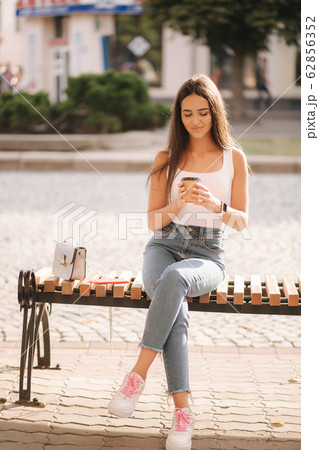 Attrective brunette sitting on the bench in centre of the city. Beautiful young woman ouside drink coffee Attrective brunette sitting on the bench in centre of the city. Beautiful young woman ouside drink coffee 62856352