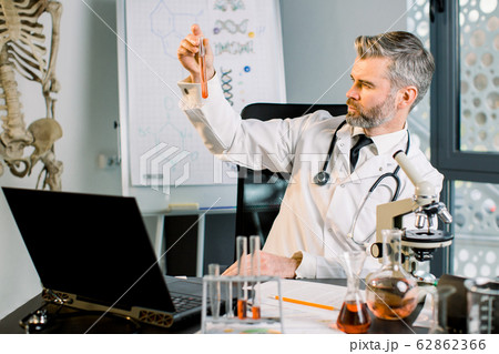 Caucasian concentrated male scientist doctor working in lab, doing research and making experiment, holding test tube with new vaccine sample or drug, looking at tube. Virology, biology, medicine Caucasian concentrated male scientist doctor working in lab, doing research and making experiment, holding test tube with new vaccine sample or drug, looking at tube. Virology, biology, medicine 62862366