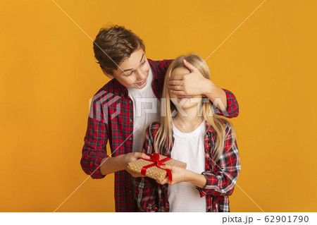 Brother Covering Sister's Eyes And Giving Birthday Gift Over Yellow Background Brother Covering Sister's Eyes And Giving Birthday Gift Over Yellow Background 62901790