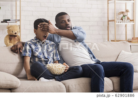 Father and son watching scary movie together, dad covering kid's eyes 62901886