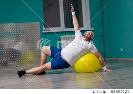 Smiling fat man is doing exercises using fitness ball. Overweight man is happy with the result of his training in group fitness classes Smiling fat man is doing exercises using fitness ball. Overweight man is happy with the result of his training in group fitness classes 62909126