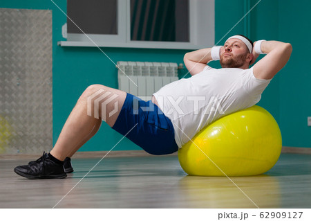 Overweight man is lying on a fitness ball during group fitness classes. Fat man looks disappointed because of bad result of weight loss trainings Overweight man is lying on a fitness ball during group fitness classes. Fat man looks disappointed because of bad result of weight loss trainings 62909127