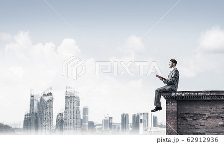 Man on roof edge reading book and cityscape at background 62912936