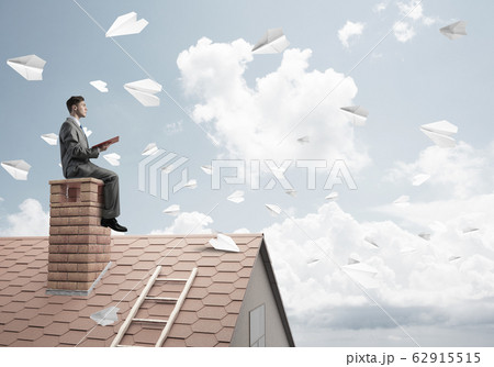 Man on brick roof reading book and paper planes flying in air Man on brick roof reading book and paper planes flying in air 62915515
