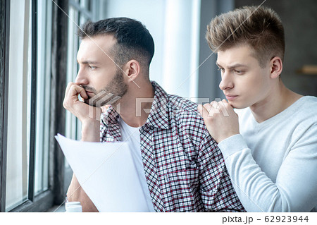 Two young men standing near the window looking desperate Two young men standing near the window looking desperate 62923944