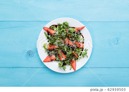 Healthy organic diet salad with arugula, strawberries and sesame with balsamic glaze in white plate on a pastel blue wooden background Healthy organic diet salad with arugula, strawberries and sesame with balsamic glaze in white plate on a pastel blue wooden background 62939503