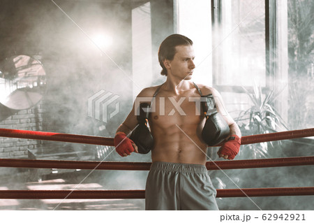 Male boxer having muscular body standing inside a boxing ring. Boxer resting his arms on the rope of boxing ring. Male boxer having muscular body standing inside a boxing ring. Boxer resting his arms on the rope of boxing ring. 62942921