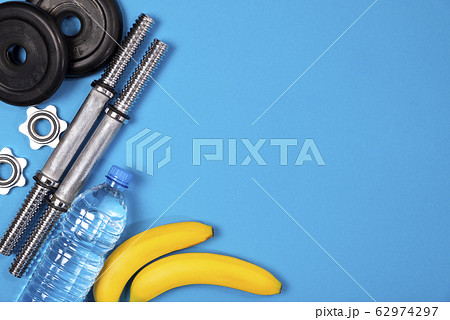 Fitness or bodybuilding. Sports equipment on a blue background, banana, water bottle, barbell, dumbbell, top view with plenty of copy space Fitness or bodybuilding. Sports equipment on a blue background, banana, water bottle, barbell, dumbbell, top view with plenty of copy space 62974297