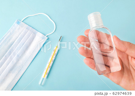 Woman hand holds antibacterial hand gel over a blue surface with surgical mask and thermometer. Woman hand holds antibacterial hand gel over a blue surface with surgical mask and thermometer. 62974639