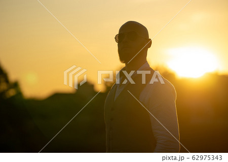 Bald Guy with a stylish beard and sunglasses on a blurred city background during dramatic sunset Bald Guy with a stylish beard and sunglasses on a blurred city background during dramatic sunset 62975343