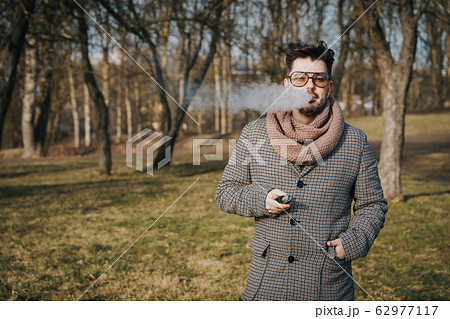 Confident young bearded male in sunglasses vape electronic cigarette. A young handsome bearded hipster man vaping e-cigarette outdoors in the park. Close up. 62977117
