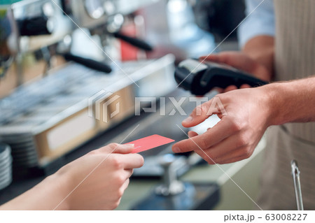 Young adult barista holding wireless terminal and taking card from client 63008227