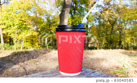 a red corrugated paper cup with coffee covered with a brown lid and a white drinking straw stands in the forest on a gray wooden table against the background of nature and trees. Copy space. 63040585