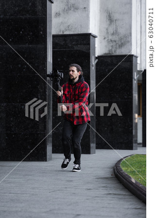 Young Professional videographer holding professional camera on 3-axis gimbal stabilizer. Pro equipment helps to make high quality video without shaking. Cameraman wearing red shirt making a videos. 63040711