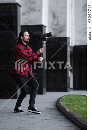 Young Professional videographer holding professional camera on 3-axis gimbal stabilizer. Pro equipment helps to make high quality video without shaking. Cameraman wearing red shirt making a videos. 63040712