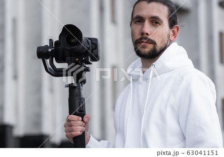 Young Professional videographer holding professional camera on 3-axis gimbal stabilizer. Pro equipment helps to make high quality video without shaking. Cameraman wearing white hoodie making a videos. 63041151