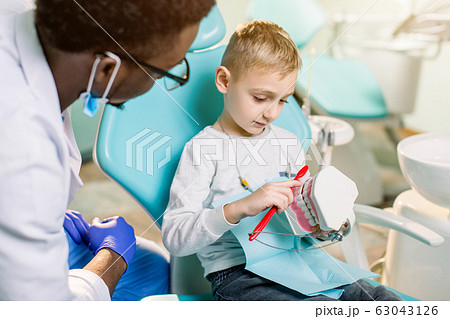 African-American ethnic black male dentist in blue latex gloves checking condition of teeth of little smiling boy. Little boy in blue dental chair in modern dentistry clinic African-American ethnic black male dentist in blue latex gloves checking condition of teeth of little smiling boy. Little boy in blue dental chair in modern dentistry clinic 63043126