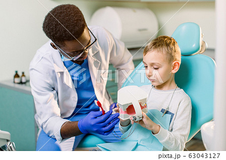 African-American ethnic black male dentist in blue latex gloves checking condition of teeth of little smiling boy. Little boy in blue dental chair in modern dentistry clinic 63043127
