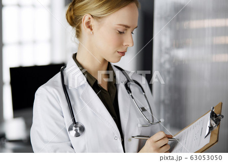 Cheerful smiling female doctor using clipboard in clinic. Portrait of friendly physician woman at work. Perfect medical service in hospital. Medicine concept 63053050