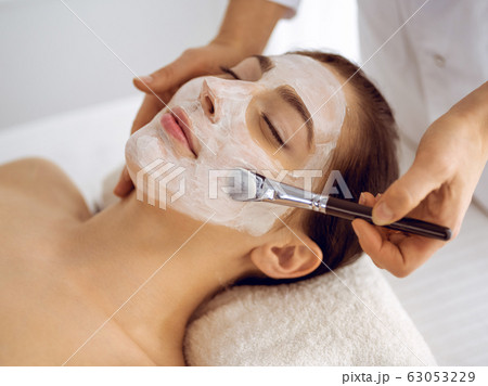 Beautiful brunette woman enjoying applying cosmetic mask with closed eyes. Relaxing treatment in medicine and spa center concepts 63053229