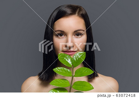 Studio beauty portrait of young brunette with natural make-up perfect skin with green exotic leaf on gray background. Concept of natural cosmetics, face and body care products. 63072418