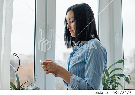 Portrait of young smiling woman in shirt at home near the window with smartphone reading text 63072472