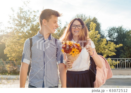 Boy and girl teenagers walking outdoor, couple smiling and talking. Sunny summer day, girl with bouquet of flowers 63072512