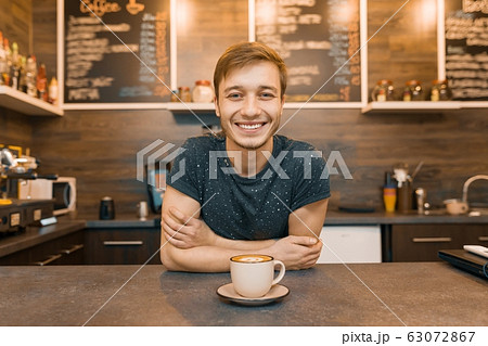 Portrait of young smiling male barista with prepared drink with arms crossed standing behind cafe counter. Coffee shop business concept Portrait of young smiling male barista with prepared drink with arms crossed standing behind cafe counter. Coffee shop business concept 63072867