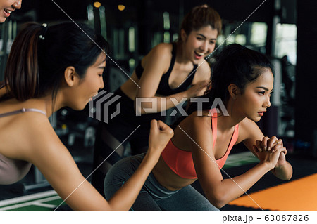 Group of people cheering on their Asian female friend doing squats with a weight plate in fitness gym. Working out together as a teamwork. 63087826