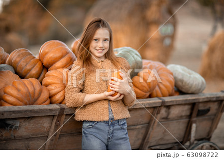 The girl stands and smiles near the cart with pumpkins and holds a small pumpkin in her hands. 63098722