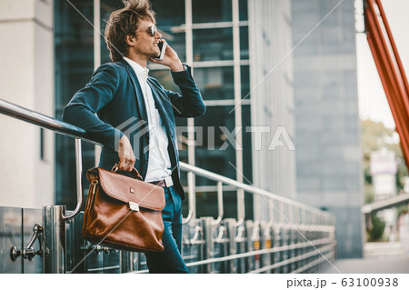 Young man in a sute standing outdoors and talking on the mobile phone and smiling Young man in a sute standing outdoors and talking on the mobile phone and smiling 63100938