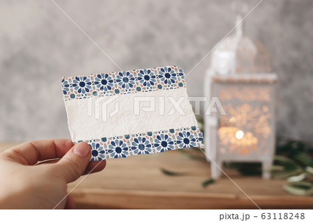 Closeup of hand holding blank paper card with watercolor Moroccan pattern. Greeting card mock-up scene. Ramadan Kareem design. Blurred background with White arabic lantern, olive leaves on table. 63118248