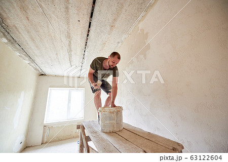 Male is making repairing in apartment by putting putty on the walls and standing on wooden stand against a window 63122604