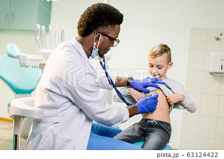Physician looking at patient and examining posture of little boy. Happy kid smiling, looking away. African American Doctor having stethoscope on neck, wearing in white medical gown 63124422